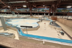 Ein großes Indoor-Wasserpark mit zahlreichen Wasserbahnen, Pfählen, Texttafeln, Deckenleuchten und Wandgemälden, das wie ein Minigolfplatz gestaltet ist.