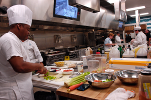 Eine Gruppe von Köchen in einer Küche, mit einem im Vordergrund in einer weißen Schürze und Mütze, der ein Messer hält und von verschiedenen Kochutensilien, Schalen, Gemüse, einem Handy, Flaschen und anderen Gegenständen umgeben ist, während im Hintergrund ein Fernseher zu sehen ist.