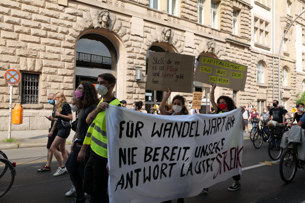 Eine Gruppe von Menschen marschiert auf einer Straße bei einer Demonstration, einige halten Schilder und andere fahren Fahrräder, im Hintergrund ein Gebäude mit Fenstern, Arkaden, Säulen und Skulpturen.