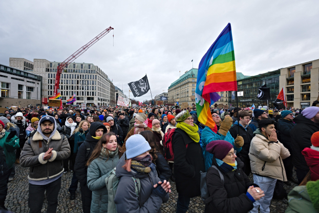 Große Gruppe von Menschen bei einer LGBTQ+-Rechte-Demonstration in Berlin, die Fahnen und Schilder mit Text halten, vor einem Gebäude mit einem Kran und einem bewölkten Himmel im Hintergrund.