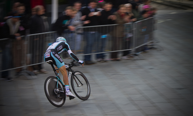 Eine Person fährt ein Fahrrad bei einem Rennen, mit Zuschauern hinter einem Zaun links.