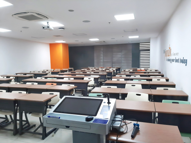 Ein Klassenzimmer mit Tischen und Stühlen in Reihen, einem deckenmontierten Projektor und Gegenständen auf den Tischen, mit Text an der rechten Wand, der darauf hinweist, dass es sich um eine Privatschule in Singapur handelt.