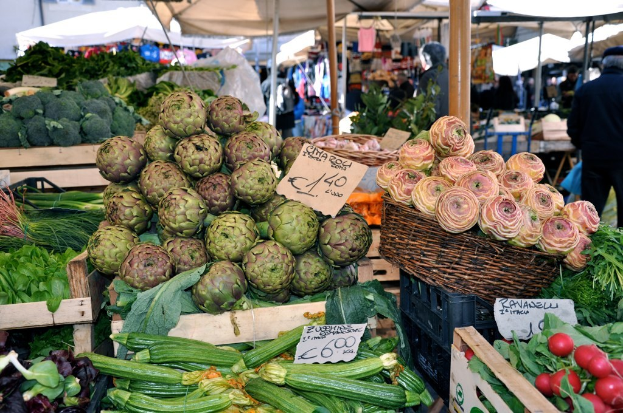 Körbe gefüllt mit Obst, Gemüse und kleinen Schildern mit Text sind unter Zelten zu sehen, mit ein paar Menschen im Hintergrund.