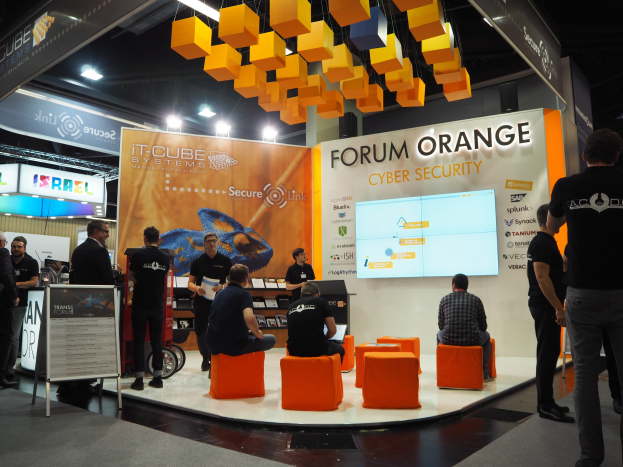 Eine Gruppe von Menschen an einem Messestand für IT-Cube Systems Forum Orange Cyber Security, mit sitzenden und stehenden Teilnehmern, einer Tafel mit Text, einem Anzeigebildschirm, Regalen mit Gegenständen, Schildern, Decke mit Leuchten und Lampen.