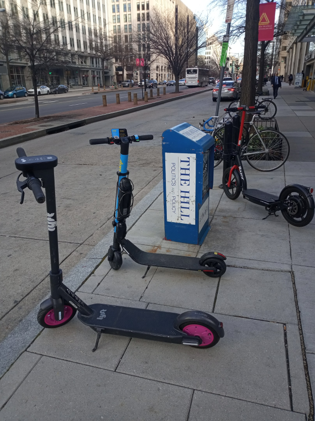 Zwei Elektro-Scooter auf einer Stadtstraße mit Fahrrädern, einem Müllcontainer, Fahrzeugen, Bäumen, Gebäuden und Menschen im Hintergrund geparkt.