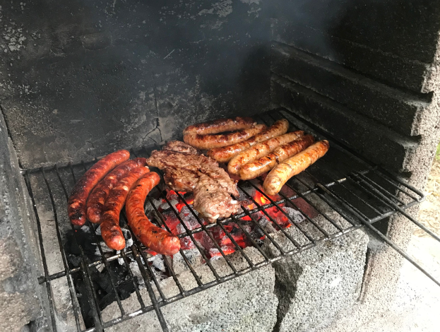 Grill mit Würstchen und Fleisch darauf, umgeben von Kohle und Feuer, draußen auf Gras.