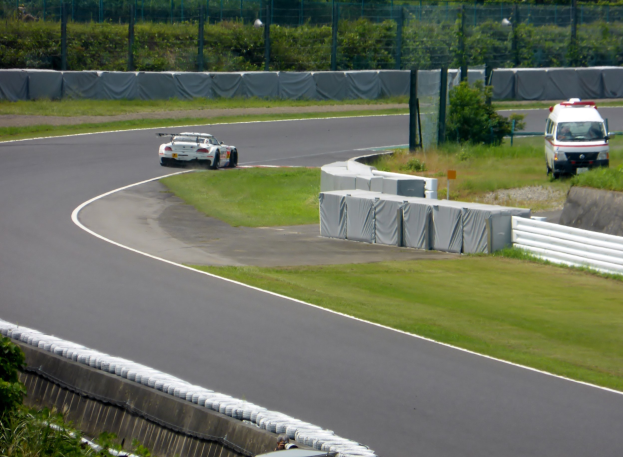 Ein Rennwagen fährt auf einer Rennstrecke mit einem Polizeiwagen im Hintergrund, umgeben von Gras, Pflanzen, Bäumen und einer Umzäunung, mit einem Fahrzeug auf der rechten Seite und Text unten.
