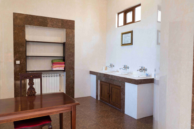 Ein Badezimmer mit einem Tisch mit Stühlen, zwei Waschbecken mit Wasserhähnen, Schränken, einem gerahmten Foto an der Wand, einem Fenster und einem Handtuchhalter.