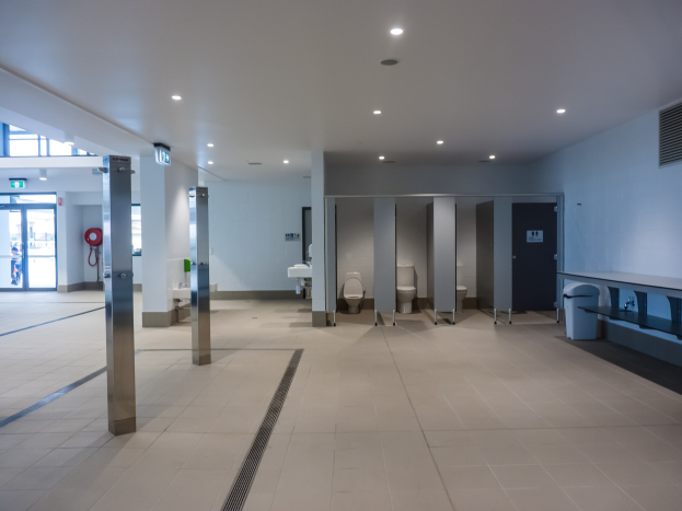 Großes öffentliches WC mit mehreren Toiletten, tragenden Säulen, einem Müllbehälter, einer Eingangstür, einer Hinweistafel, einem Feuerlöscher und Deckenleuchten.