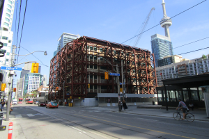 Eine belebte Stadtstraße mit einem großen Gebäude im Hintergrund, das gebaut wird, Fahrzeuge auf der Straße, ein Fahrradfahrer, Fußgänger auf dem Gehweg, Verkehrsampeln, Pfosten mit angebrachten Schildern, ein Kran und der Himmel.