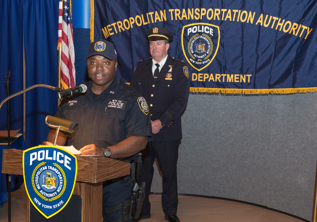 Zwei Polizeibeamte stehen vor einem Rednerpult, einer hält Papiere und spricht in ein Mikrofon, mit einer Tafel, Fahne und einem Tisch in der Nähe und einer Tafel mit Symbolen und Text im Hintergrund.