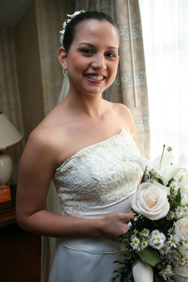 Eine Braut steht strahlend da und hält einen Blumenstrauß, mit einem Vorhang und einer Wand im Hintergrund und einer Lampe auf einem Tisch links.