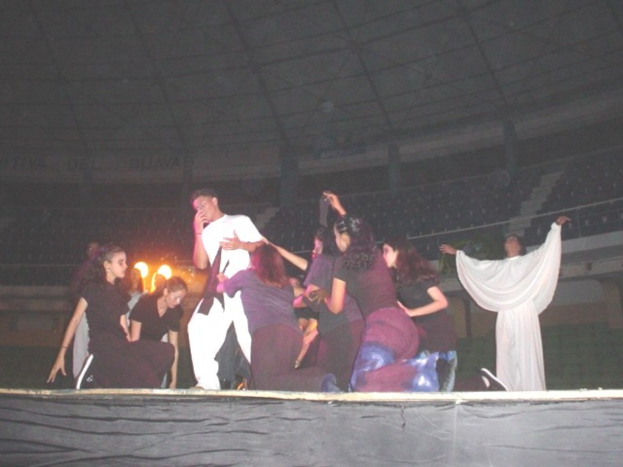 Eine Gruppe performs auf einer Bühne mit Sitzplätzen, Grills und einem Schuppen im Hintergrund.