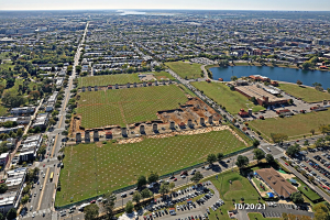 Luftaufnahme einer Stadt mit Gebäuden, Bäumen, Gras, Straßen mit Fahrzeugen, Wasser und Wolken, die einen großen zentralen Bereich als Parkplatz zeigt.