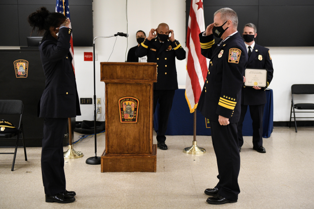 Eine Gruppe uniformierter Personen in Masken steht vor einem Podium mit einem Mikrofon, umgeben von Flaggen, Stühlen und Tischen, während eine neue Feuerwehrchef vereidigt wird, wobei eine Person ein Zertifikat hält.