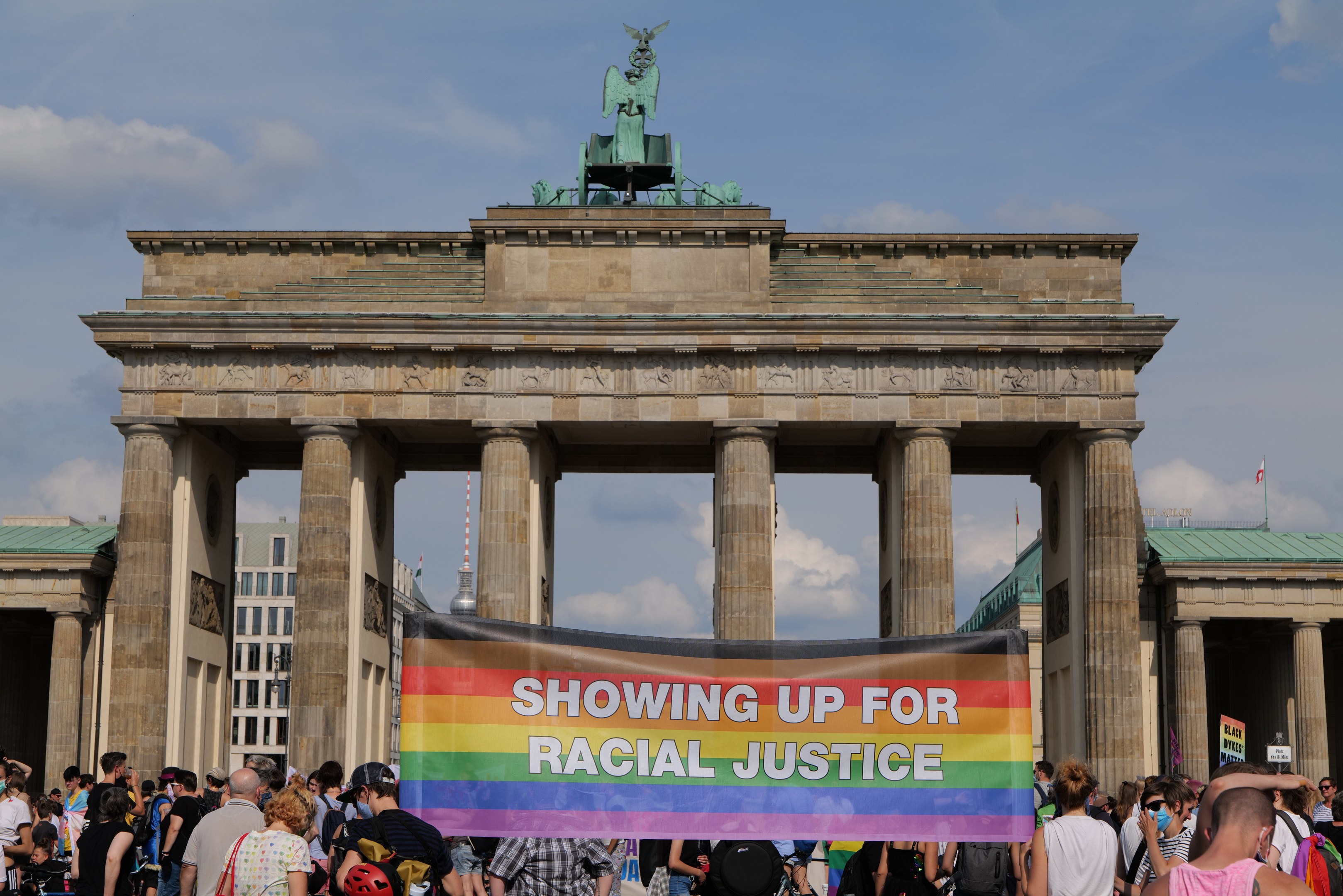 Eine Gruppe von Menschen mit einer "Racial Justice"-Schriftzug-Tafel vor dem Brandenburger Tor in Berlin, Deutschland, mit Gebäuden und bewölktem Himmel im Hintergrund.