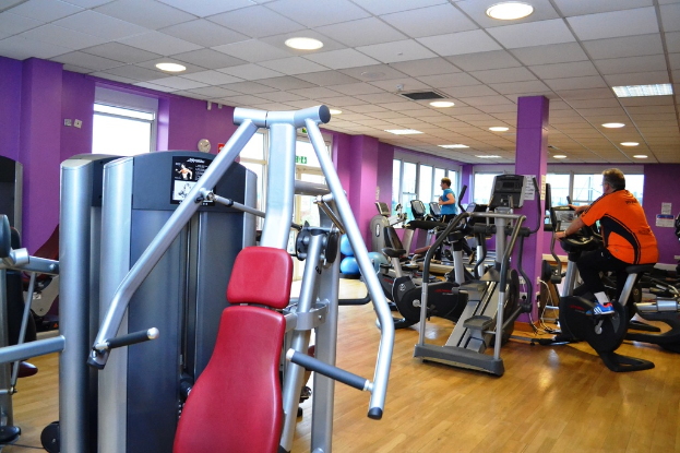 Ein Fitnessstudio mit verschiedenen Geräten und einigen Menschen, die trainieren, mit einer Decke und Lampen oben im Bild.