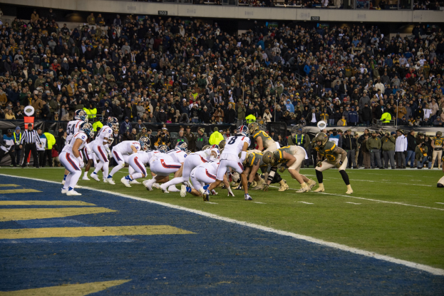 Ein Fußballspiel im Gange mit Spielern in Sportkleidung und Helmen auf dem Boden, von einem Stadionplatz aus mit Zuschauern und Beschilderung im Hintergrund betrachtet.