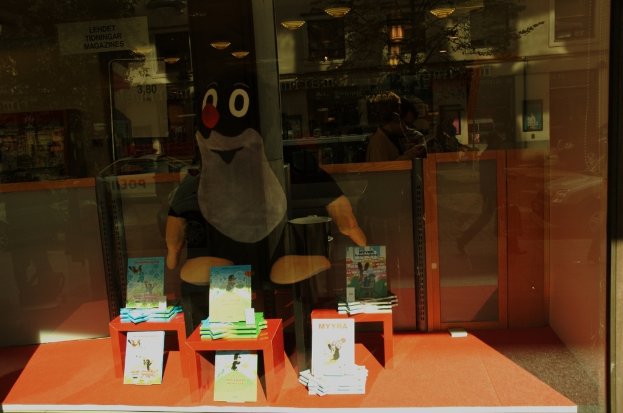 Ein Clown steht in der Mitte des Bildes, mit einem Tisch und Karten unten und Spiegelungen von Menschen auf dem Glas.