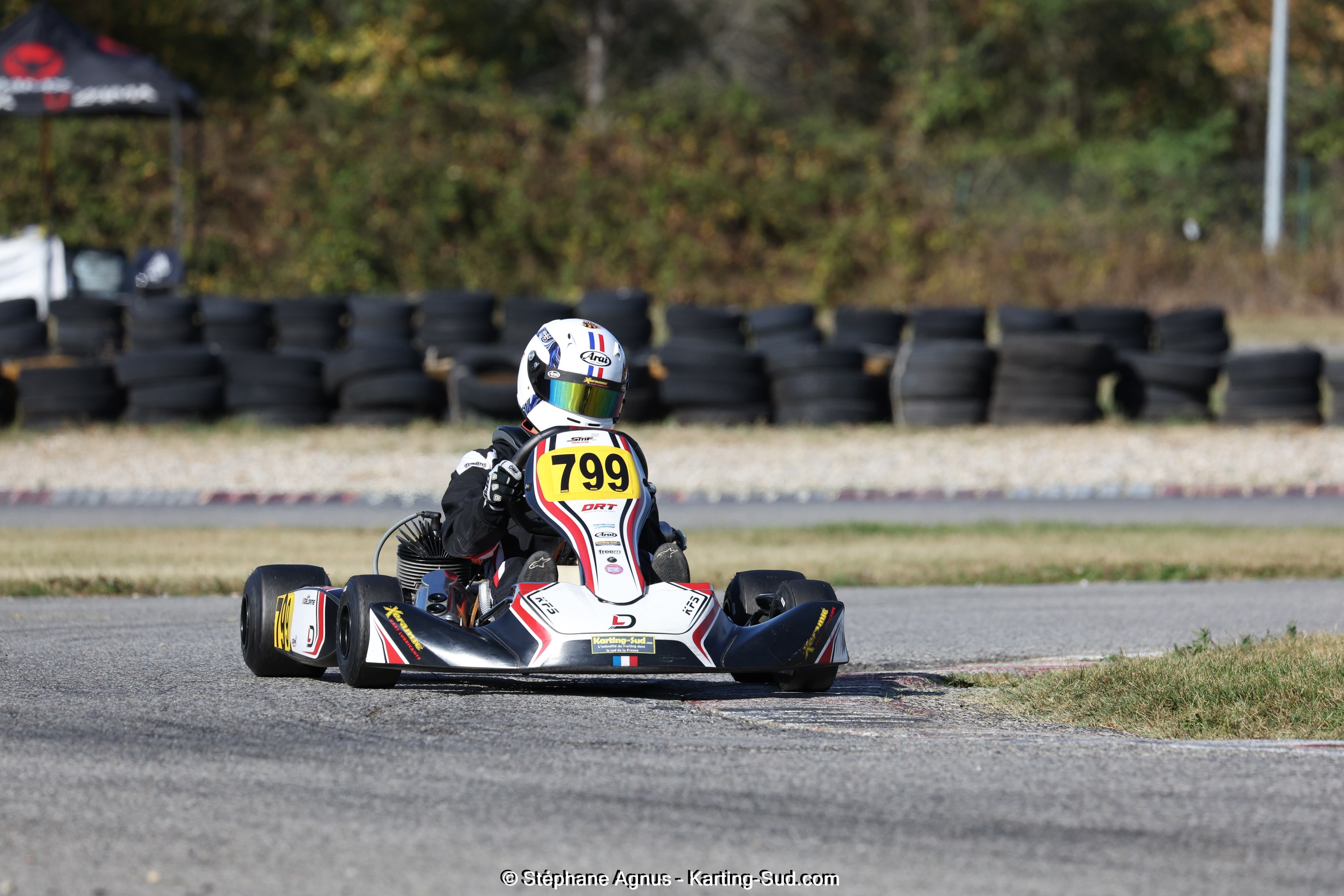 Eine Person in einem Helm fährt ein Go-Kart auf einer Rennstrecke, umgeben von Gras, mit Reifen, einem Zelt, einem Pfahl und Bäumen im Hintergrund und Text unten.