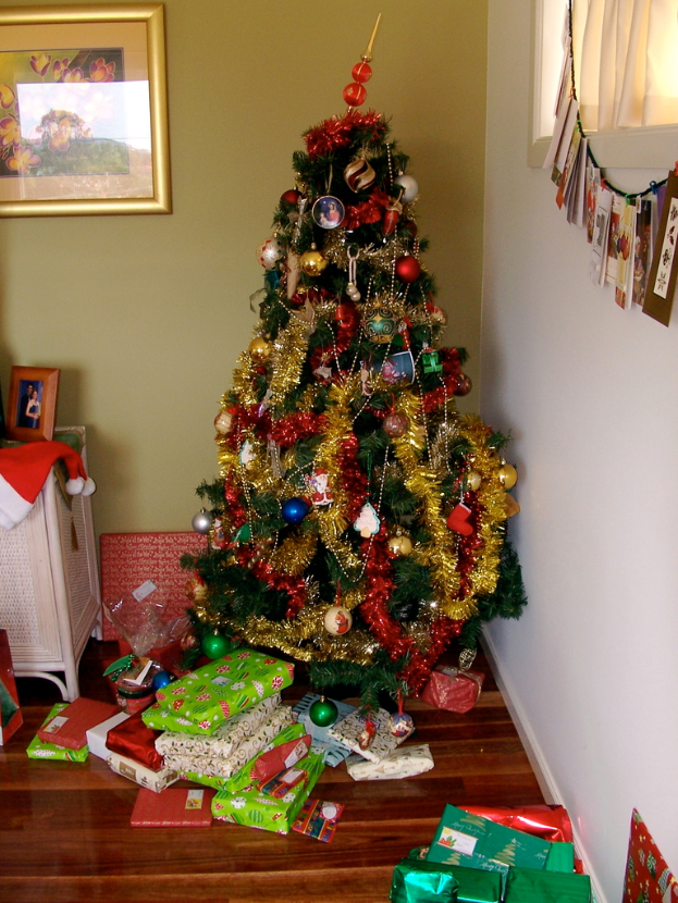 Ein geschmückter Weihnachtsbaum mit Geschenken am Boden, ein Foto auf der linken Wand und ein Vorhang am rechten Fenster.