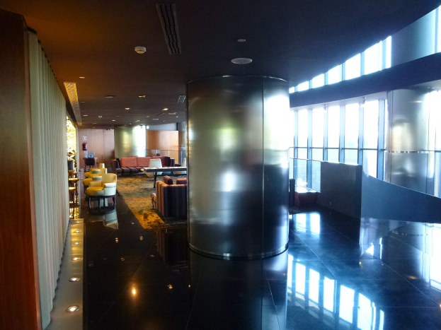 Geräumige First-Class-Lounge am Hong Kong International Airport mit großen Fenstern, hohen Decken mit elektrischen Leuchten, Sitzanordnungen einschließlich Stühle und Sofas, Tischen, einem Teppichboden und einer zentralen Säule.