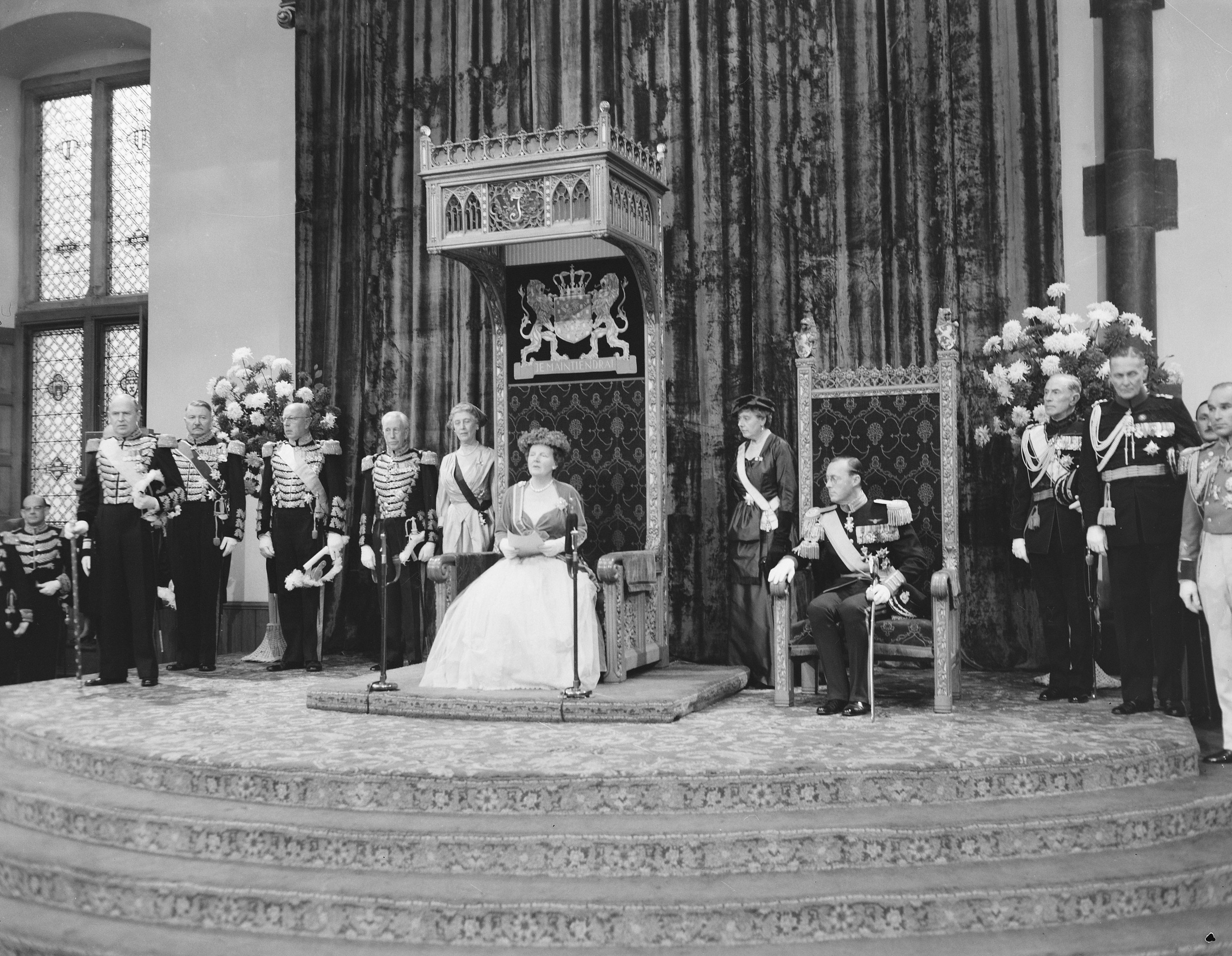 Schwarz-weiß-Foto der Queen Elizabeth II. sitzend in einem Stuhl in einem weißen Hochzeitskleid und mit einem Bukett, mit Prinz Philip zu ihrer Rechten und Begleitern zu ihrer Linken.