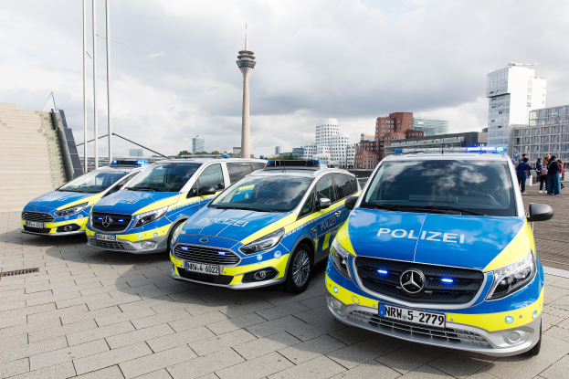 Drei Mercedes-Benz V-Klasse Polizeifahrzeuge nebeneinander vor einem Gebäude geparkt, mit einer Gruppe von Menschen rechts stehend und einer Stadtlandschaft mit Gebäuden, Masten, einem Turm, Bäumen und einem klaren blauen Himmel im Hintergrund.