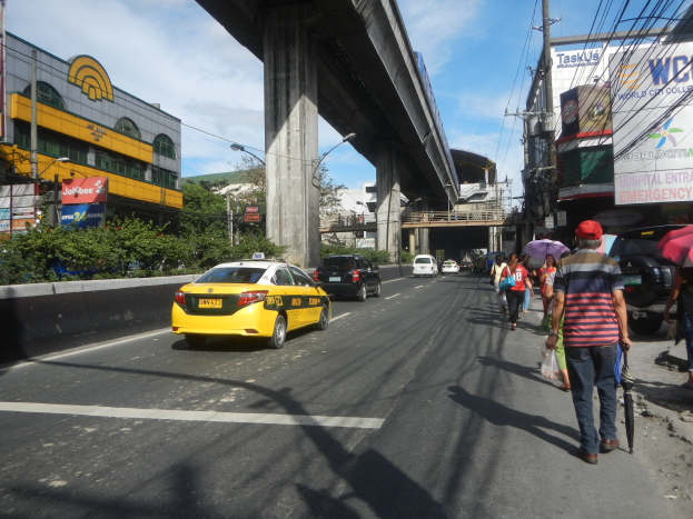 Gelbes Taxi fährt auf einer Straße mit hohen Gebäuden, Menschen auf dem Gehweg mit Regenschirmen, eine Brücke im Hintergrund, bewölkter Himmel und Strommasten mit Drähten entlang der Straße.