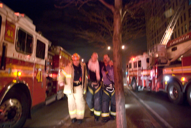 Drei Personen vor einem Feuerwehrauto auf der linken Seite und einem Gebäude auf der rechten Seite mit einem Baum in der Mitte.