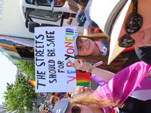 Eine Gruppe von Menschen hält ein Schild mit der Aufschrift "Die Straßen sollten sicher sein" vor einem Lastwagen, einige tragen Mützen und Schutzbrillen, andere Schilder, vor einem Hintergrund aus Bäumen, Gebäuden und einem klaren blauen Himmel.