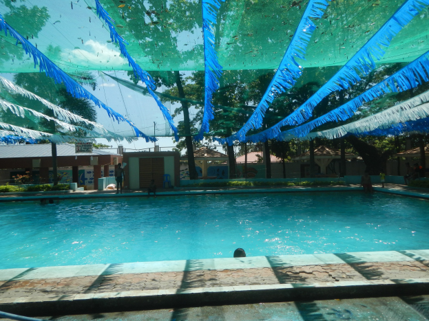 Ein großes Schwimmbecken mit blauen und grünen Bändern an der Decke, umgeben von Häusern, Bäumen und einem klaren blauen Himmel, mit Menschen, die das Wasser und die Sonne genießen.