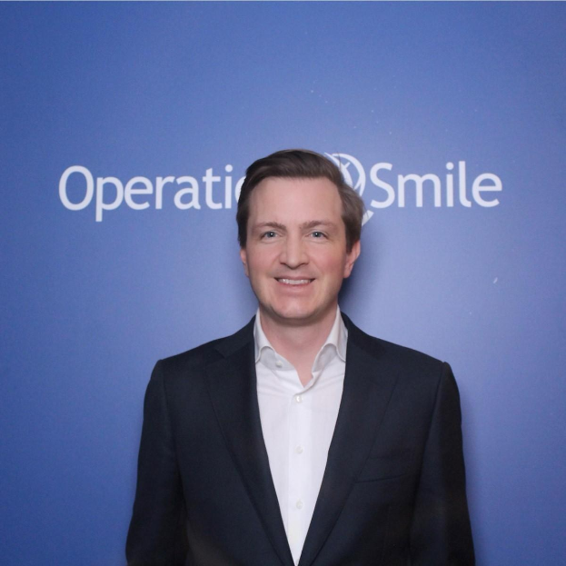 Ein Mann in einem Anzug steht vor einer blauen Wand mit der Aufschrift "Operationsmile" und lächelt