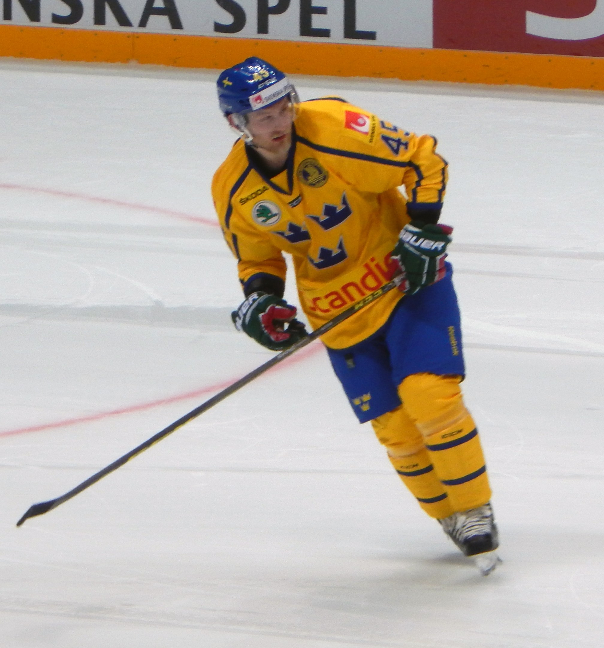Eishockeyspieler in Bewegung auf dem Eis, mit Helm, Handschuhen und Schlittschuhen, einen Hockeystock haltend, mit einer Tafel im Hintergrund mit Text.