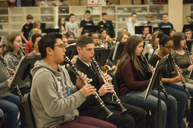 Eine Gruppe von Menschen, die Klarinetten in einem Klassenzimmer spielen, einige sitzen mit Notenpulten, andere stehen, mit Regalen voller Kisten im Hintergrund.