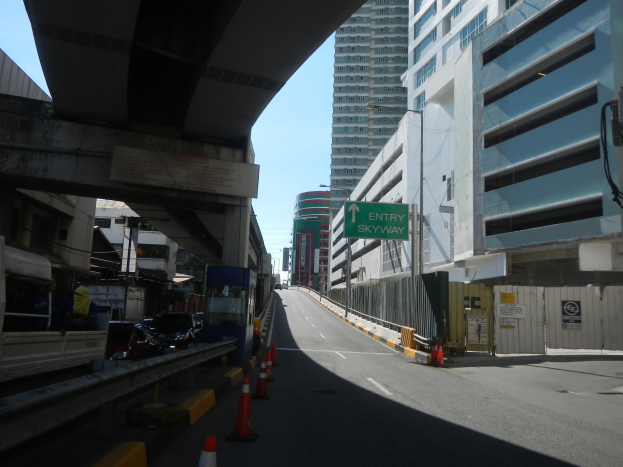 Stadtstraße mit Verkehrskegeln, Fahrzeugen, einer Brücke auf der linken Seite, Gebäuden mit Namensschildern, Laternen und einem Himmel als Hintergrund.