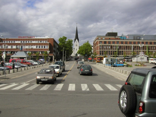 Stadtstraße mit parkenden Autos, Gebäuden, Bäumen, Laternen, wolkenverhangenem Himmel und sichtbarem Kfz-Kennzeichen.