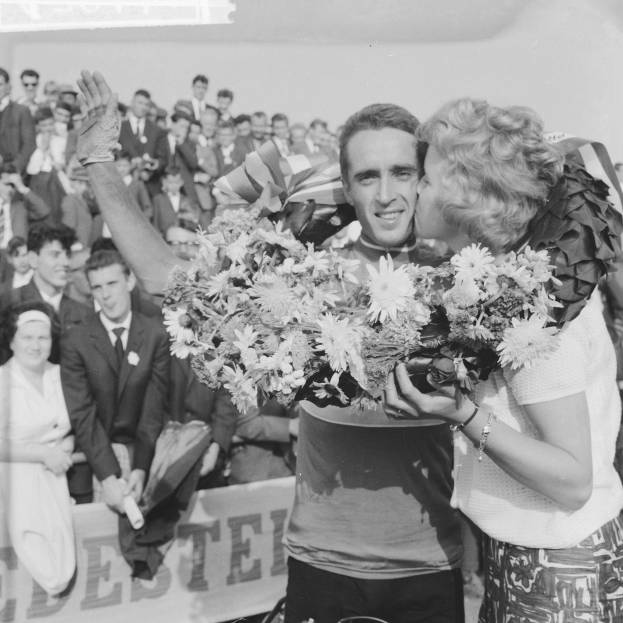 Ein Schwarz-Weiß-Foto eines Mannes und einer Frau, die sich vor einer Menge umarmen und küssen, wobei der Mann einen Blumenstrauß hält und die Frau lächelt, während die Menschen hinter ihnen klatschen.