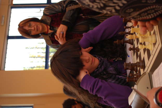 Ein Mädchen in einem lila T-Shirt sitzt beim Schachspielen, während eine Frau in einer Jacke hinter ihr steht, vor einem gelben Hintergrund mit einer Glastür.