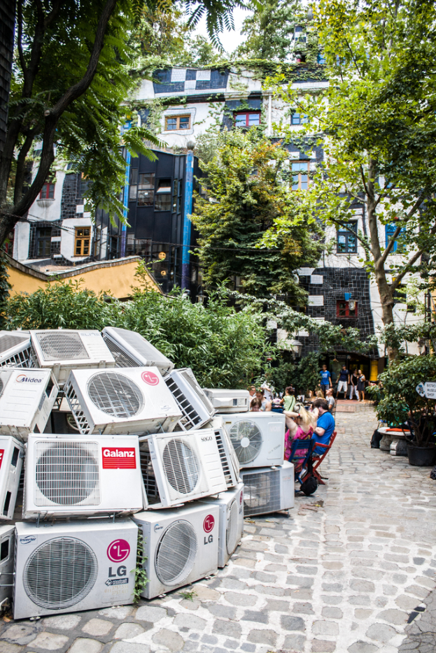 Luftkühler am Boden links unten, ein Weg nach rechts, Menschen auf Stühlen neben dem Weg, mit Gebäuden, Bäumen und Pflanzen oben.