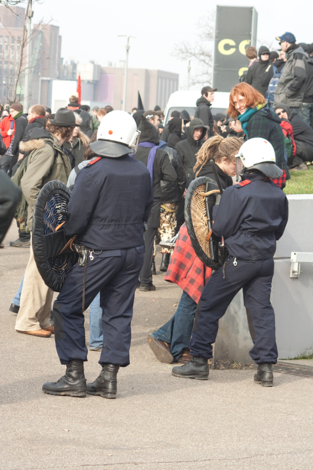 Eine Gruppe von Menschen, die auf einer Straße gehen, mit zwei Personen vorne, die wie Polizisten aussehen, Gebäude im Hintergrund und Boden unten.