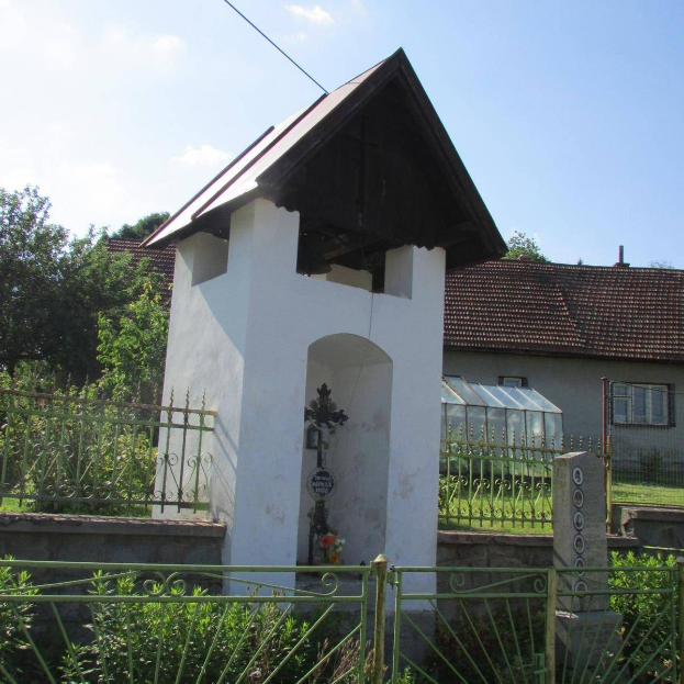 Kleine weiße Kirche mit einem Kreuz darauf, umgeben von einem Metallzaun, Pflanzen und einem Weg, vor Häusern und einem bewölkten Himmel, als die Kirche des Heiligen Grabes identifiziert.