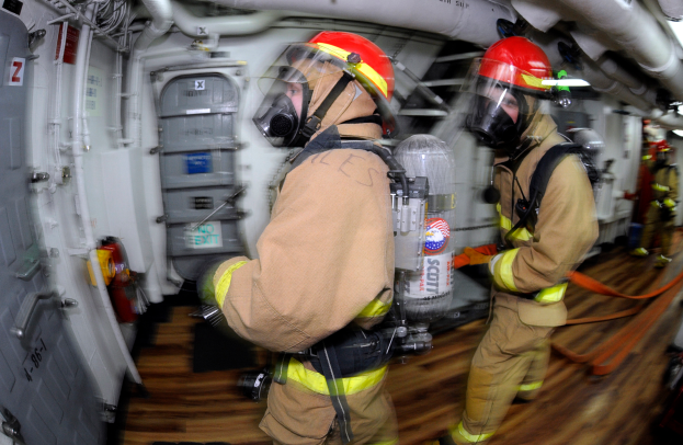 Zwei Feuerwehrleute in Schutzausrüstung, einschließlich Helmen und Gasmasken, gehen durch das Innere eines Flugzeugs mit einer Tür auf der linken Seite und Rohren und anderen Gegenständen auf der rechten Seite.
