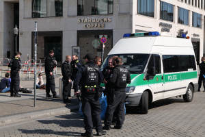Eine Gruppe von Polizisten steht vor einem Starbucks-Café, rechts daneben parkt ein Van, links stehen und sitzen Menschen, im Hintergrund ein Gebäude mit Fenstern, einer Tafel, einem Laternenmast und einem Zaun.