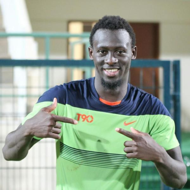 Ein Mann in einem grünen und blauen Hemd, der in die Kamera lächelt und zeigt, mit einem leicht unscharfen Geländer, Wand und Tür im Hintergrund.