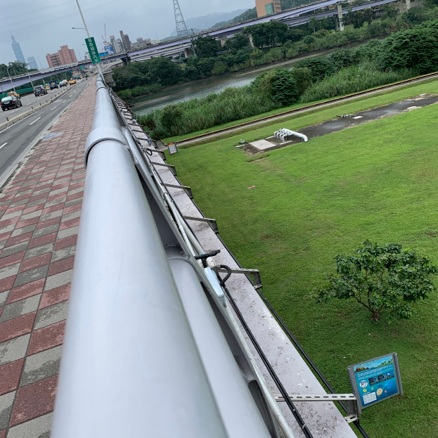 Eine Stadtansicht von einem Gebäude aus, mit einem prominenten Metallwasserrohr im Vordergrund, sowie Straßen, Fahrzeuge, Grünflächen und verschiedene Strukturen im Hintergrund unter dem Himmel.