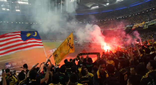 Eine große Menge Menschen in einem Stadion mit Bengalos, Fahnen und einer Kamera, mit Rauch und Schildern mit der Aufschrift "Malaysia vs Malaysia Live-Streaming" im Hintergrund.