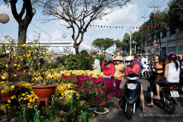 Eine Gruppe von Menschen in Helmen und Mützen, einige gehen und einige fahren Fahrräder auf einer Straße, mit Pflanzen, einem Blumentopf auf einem Hocker, Bäumen, einem Pfahl, Drähten, einer Lampe, einer Brücke und Gebäuden in der Nähe.