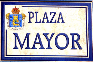 Rechteckige weiße Plakette mit schwarzem Text "Plaza Mayor" in fetter Schrift, blauem Rahmen und einem gekrönten Wappen-Logo an einer Gebäudefassade.