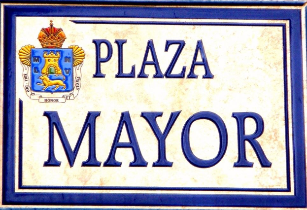 Rechteckige weiße Plakette mit schwarzem Text "Plaza Mayor" in fetter Schrift, blauem Rahmen und einem gekrönten Wappen-Logo an einer Gebäudefassade.